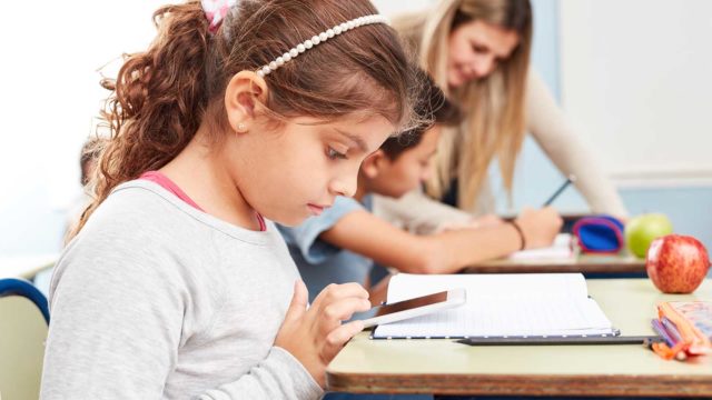Foto: Shutterstock. Jente sitter med mobiltelefon ved pulten i klasserommet. Lærer i bakgrunnen.