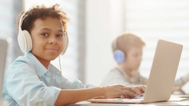 Foto: Shutterstock / AnnaStills. Bildet viser en gutt med datamaskin og hodetelefoner på skolen.