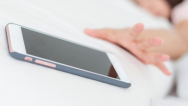 Foto: Shutterstock / leungchopan. Bildet vsier en telefon på en hvit pute, en hånd strekker seg etter telefonen.